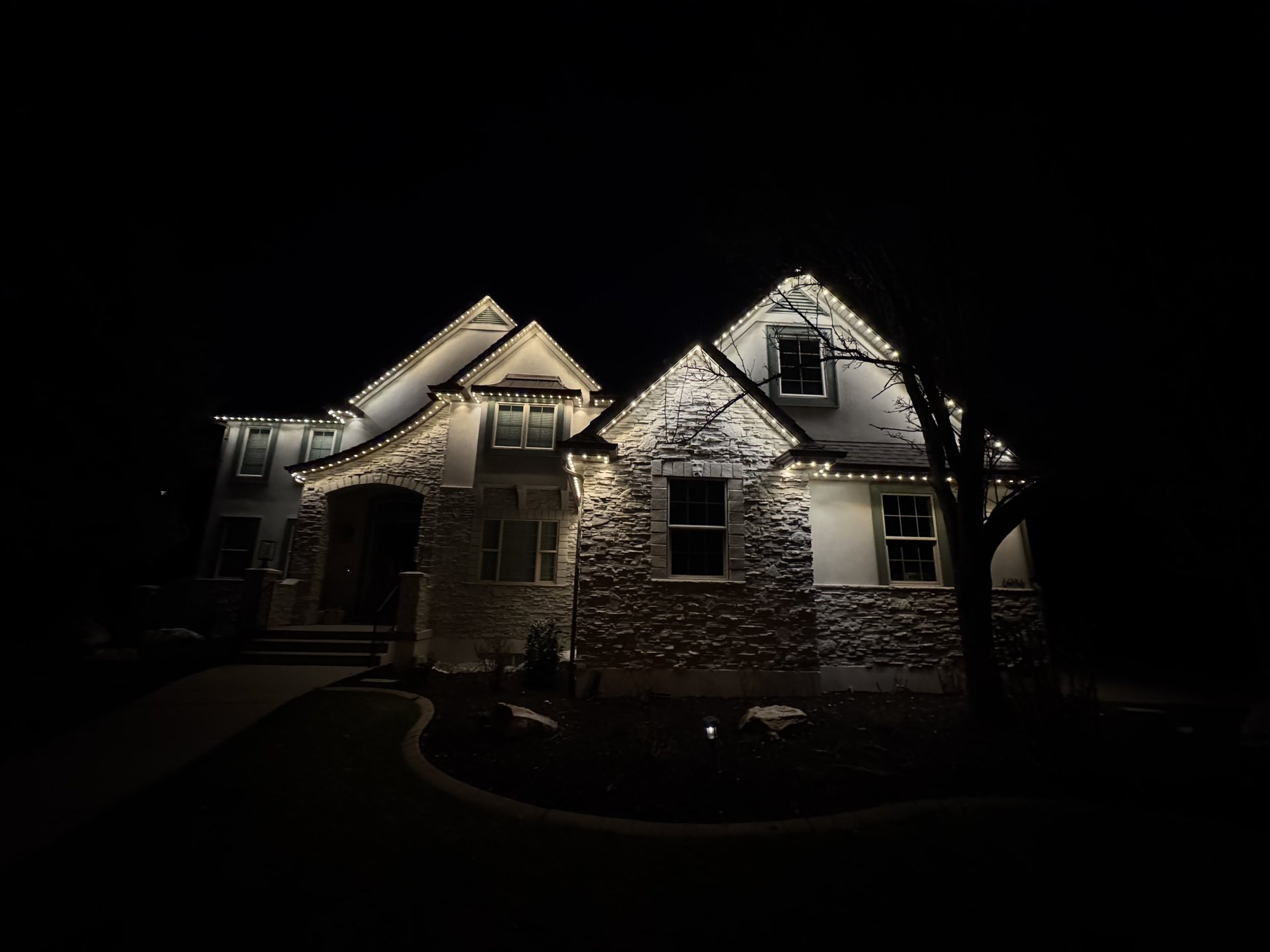 Warm white permanent lighting outlining the roofline of a two-story stone home at night