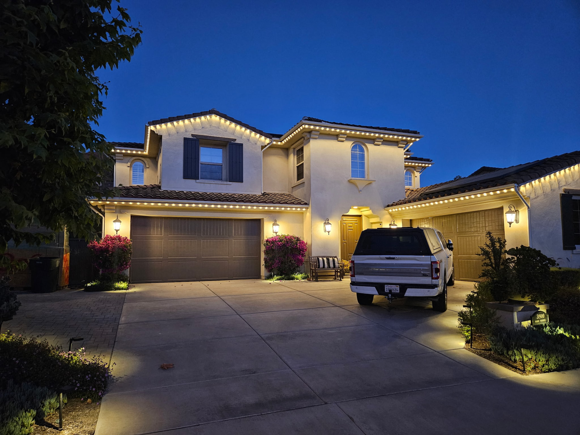 Warm white permanent lighting on a two-story Spanish-style home at dusk